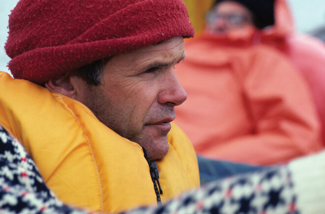 The image shows a man wearing a red hat and a yellow life vest. He has a serious expression on his face and is looking off to the side. In the background, another person wearing an orange jacket is partially visible. The overall impression is one of a potentially cold or outdoor environment.